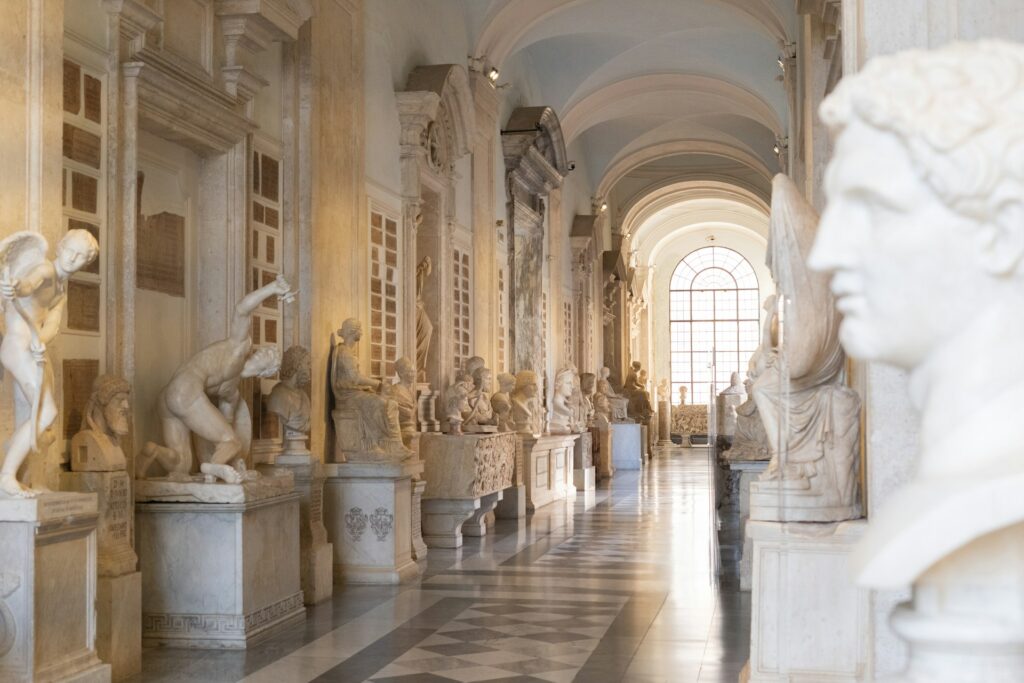 A long corridor in a grand building, brightly lit by a large window and filled with Roman statues.